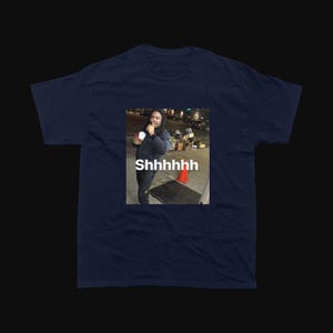 Op de afbeelding: Marineblauw T-shirt met een meme van een persoon die een beker vasthoudt en het woord "Shhhhhhh" op de afbeelding. De afbeelding is vierkant en gecentreerd op het shirt.