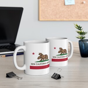 May include: Two white ceramic mugs with the New California Republic emblem. The design features two brown bears, a red star, and the text "NEW CALIFORNIA REPUBLIC" above a red stripe. The mugs are on a light-colored surface.