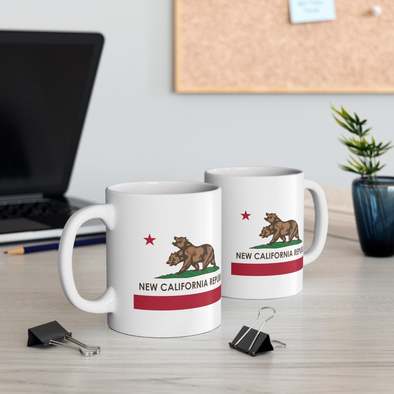 May include: Two white ceramic mugs with the New California Republic emblem. The design features two brown bears, a red star, and the text "NEW CALIFORNIA REPUBLIC" above a red stripe. The mugs are on a light-colored surface.