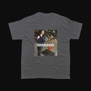 Op de afbeelding: Donkergrijs t-shirt met een afgedrukte afbeelding van een persoon die een beker vasthoudt, met het woord "Shhhhhhh" in witte tekst. Het ontwerp is een meme, geschikt voor vrijetijdskleding.