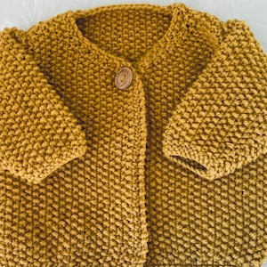 Könnte beinhalten: Ein senfgelber Strickcardigan mit Knopfleiste. Der Cardigan ist mit einem strukturierten Stich gearbeitet.