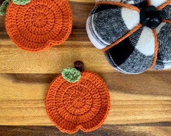 May include: Two crocheted pumpkin coasters in orange with green and brown leaves. The coasters are on a wooden surface next to a larger white and black plaid pumpkin.