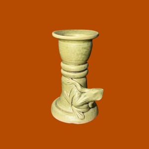 May include: A pale green ceramic candle holder featuring a sculpted frog relief on the base. The cylindrical stand has a flared top rim and a wide, stable circular foot, with a smooth, matte finish and visible surface texture throughout the piece.