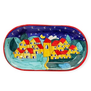 May include: An oval ceramic tray featuring a hand-painted scene of a village with yellow houses and red roofs under a dark blue night sky with a crescent moon and white stars. The tray has a distinct red border around the entire outer edge.