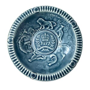 May include: A blue ceramic commemorative plate with a raised relief design. The plate features a central emblem with a crown and the text "1977 The Queen's Silver Jubilee." The plate's rim has a ridged pattern. Various animals are depicted around the central emblem.