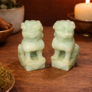 Może przedstawiać: Dwie jadeitowe statuetki Foo Dog. Każda statuetka jest szczegółowo wyrzeźbiona, przedstawiając lwa strażnika z groźną miną. Statuetki są osadzone na kwadratowych podstawach i eksponowane na drewnianej powierzchni.