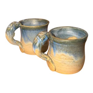 Peut inclure: Deux mugs en céramique faits à la main avec des anses uniques. Les mugs présentent un mélange de glaçures bleues, beiges et vertes. Les anses sont sculptées avec un motif texturé. Parfaits pour le café ou le thé.