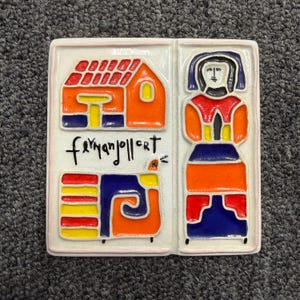 May include: A ceramic tile with a colorful, abstract design. The tile is divided into two sections, featuring a house, a figure, and a stylized animal. The design incorporates orange, red, yellow, blue, and black, with the artist's signature in the center.