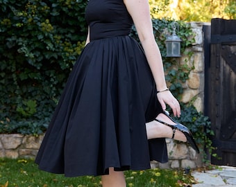May include: A black dress with a fitted bodice and a flared skirt. The dress has short sleeves and a defined waist. The person is wearing black, pointed-toe slingback heels. The background includes greenery and a stone wall.