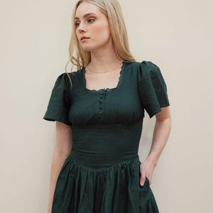 May include: A dark forest green midi dress featuring a square neckline with delicate lace trim, short puff sleeves, and a fitted bodice with a vertical row of small matching buttons. The skirt is gathered at the waist and includes side pockets.