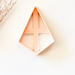 May include: A light peach-colored diamond-shaped cookie cutter with a cross-shaped interior design. The cutter is made of plastic and is set against a white background. The cutter is likely used for baking or crafting.