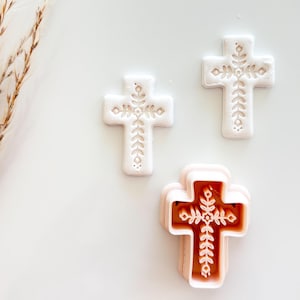 May include: Three white cross-shaped cookie cutters with floral designs. One cutter is a stack of pink and white layers. The crosses have detailed leaf and floral patterns, perfect for baking religious-themed cookies or treats.