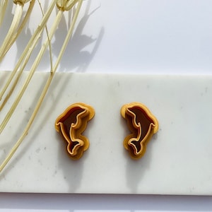 May include: A pair of dolphin-shaped stud earrings. The earrings feature a layered design with a light brown outer layer and a dark brown inner layer. The earrings are displayed on a white marble surface.