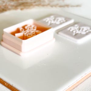 May include: A light pink floral stamp and two floral impressions on a white surface. The stamp is rectangular with a floral design inside. The impressions are square and also feature the floral design. The background is white.