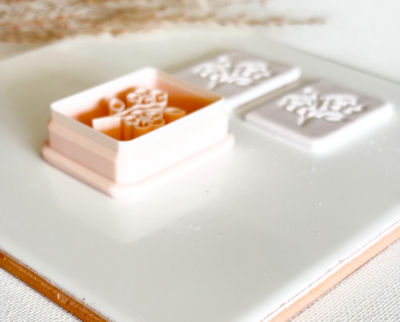 May include: A light pink floral stamp and two floral impressions on a white surface. The stamp is rectangular with a floral design inside. The impressions are square and also feature the floral design. The background is white.
