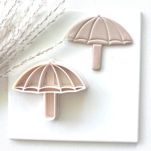 May include: Two light pink umbrella-shaped cookie cutters. One cutter is a solid umbrella shape, and the other has a detailed design with ribs. The cutters are on a white surface, with a sprig of dried plants in the upper left corner.