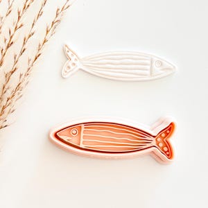 May include: Two fish-shaped cookie cutters. One is white, the other is salmon-colored. Both have detailed lines to represent the fish's scales and fins. The cutters are designed for baking or crafting, adding a nautical theme.