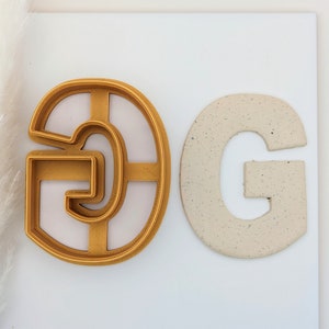 May include: A gold plastic cookie cutter in the shape of the letter G, next to a beige ceramic letter G.