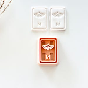 May include: Three rectangular cookie cutters. Two are white with an eye design and the letter "N". The third is a pinkish-brown color with an eye design and the letter "И".