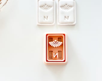 May include: Three rectangular cookie cutters. Two are white with an eye design and the letter "N". The third is a pinkish-brown colour with an eye design and the letter "И".