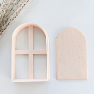 May include: A set of two pink plastic cookie cutters shaped like an arched window with four panes. The cutter on the left is the outline of the window, and the cutter on the right is the inner portion of the window.