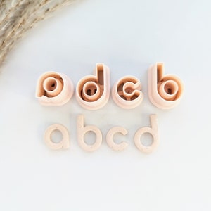 May include: Set of eight pink plastic alphabet cookie cutters. The cutters are in the shape of the letters a, b, c, and d. There are two of each letter, one with a raised design and one with a flat design.