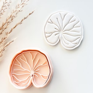 May include: Two leaf-shaped cookie cutters. One is white, the other is light pink. Both have detailed vein patterns. The cutters are on a white surface, with dried ornamental grass in the background. These are ideal for baking and crafting.