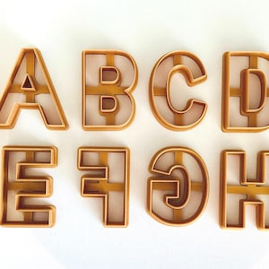 May include: A set of eight gold-colored cookie cutters shaped like the letters A through H. The cutters are made of metal and have a smooth, shiny finish.