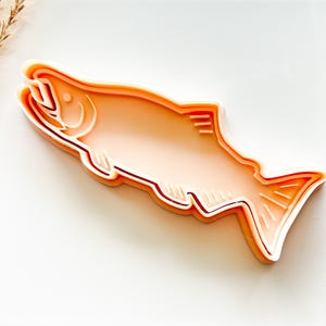May include: A salmon-shaped cookie cutter in a light peach color with white accent lines. The cutter has a detailed outline of the fish, including gills, fins, and scales. The design is layered, giving it a three-dimensional effect.