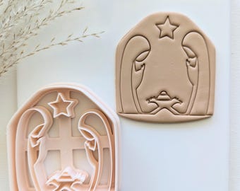 May include: A set of two pink plastic cookie cutters shaped like a nativity scene. The cutters are designed to create a manger with a star above and two figures inside.