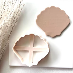 May include: A light pink seashell-shaped cookie cutter and a seashell-shaped piece of clay. The cookie cutter has a cross-shaped interior design. The clay is a solid, smooth shape. The items are on a white surface.