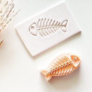 May include: A fish-shaped stamp and its impression. The stamp is a rectangular block with a fish skeleton design. The impression is a fish skeleton in orange with white outlines. The items are on a white surface.