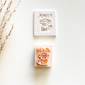 May include: Two square stamps, one white and one peach-colored, each featuring a monkey design and the word "JOKER". The white stamp has a raised design, while the peach stamp has an etched design. The stamps are likely for crafts or card making.