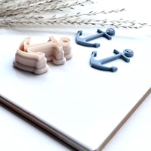 May include: Two anchor-shaped cookie cutters, one pink and one blue. The pink cutter is larger and has a more detailed design. The blue cutter is smaller and simpler. Both cutters have a heart-shaped detail at the top.