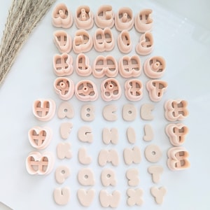 May include: A set of 26 pink plastic alphabet cookie cutters. The cutters are arranged in alphabetical order, with the letter "A" at the top left and the letter "Z" at the bottom right. The cutters are all the same size and shape, and they have a smooth, rounded edge.