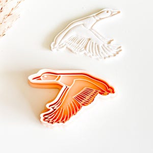 May include: Two duck-shaped cookie cutters. One is white, showing the duck in flight with detailed wing feathers. The other is orange, with a similar design, showing the duck in flight. The cutters are likely for baking or crafting.