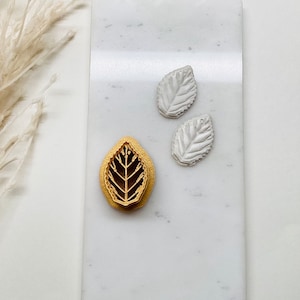 May include: Three leaf-shaped molds on a white marble surface. Two of the molds are white and one is gold.