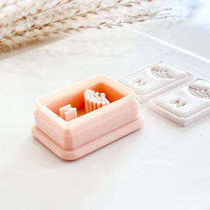 May include: A light pink rectangular cookie cutter with a small design inside, next to two rectangular molds with a face design. The items are on a white surface with a sprig of dried grass in the background.