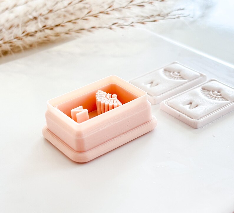 May include: A light pink rectangular cookie cutter with a small design inside, next to two rectangular molds with a face design. The items are on a white surface with a sprig of dried grass in the background.
