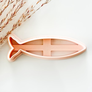 May include: A light peach-colored plastic cookie cutter shaped like a fish with a cross symbol integrated into the center. The cutter features a raised edge for cutting and a central cross-shaped structure for imprinting details into dough.