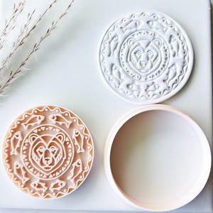 May include: A set of three round cookie cutters. Two cutters feature a bear design surrounded by fish, and the third is a plain circle. The bear design is carved into the cutters, creating a detailed relief. The cutters are light brown and white.