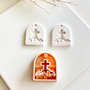 May include: Three white, arched-shaped clay pieces with a cross and mountain design, and a cross-shaped cookie cutter with a cross and mountain design. The cookie cutter is orange and red. The items are on a white surface.