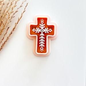 May include: A decorative cross-shaped cookie cutter. The cutter features a detailed design with a white floral pattern on a brown background, framed by a light pink border. The cross is set against a white backdrop.