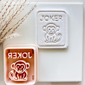 May include: A light pink cookie cutter and a white embossed design of a joker card. The card features a monkey illustration and the word "JOKER". The cookie cutter is in the shape of the card, with the word "JOKER" in reverse.