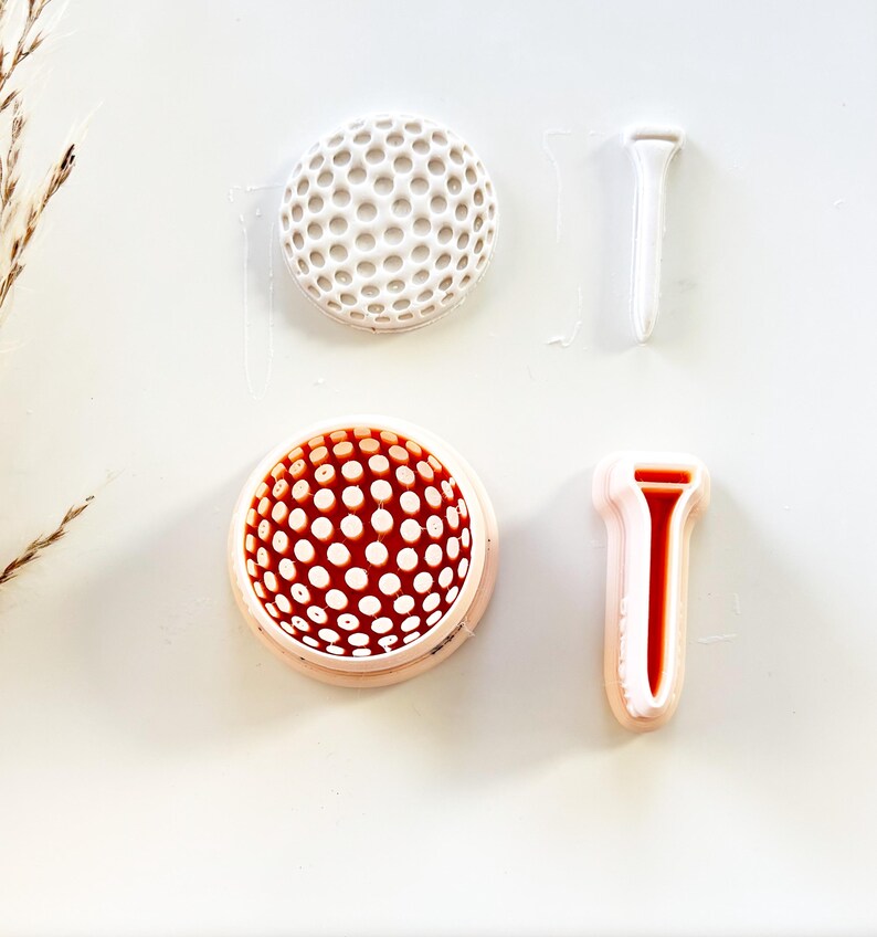 Pu&ograve; includere: Due stampi per palline da golf, uno bianco e uno rosso, con tee da golf bianchi e rossi abbinati. Le palline da golf hanno una superficie a fossette. I tee da golf sono a forma di T. Gli oggetti sono su sfondo bianco.