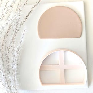 May include: A light beige semi-circular clay shape and a matching light pink semi-circular cutter with a cross-shaped interior frame. The cutter features a flat base and a curved top edge, designed for creating precise shapes in crafting materials.