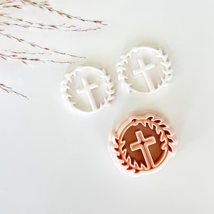 May include: Three cookie cutters with a cross design. Two are white, featuring a cross within a wreath. The third is a two-tone design, with a brown cross and wreath inside a pink frame. Ideal for baking.