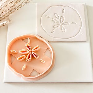 May include: A light peach-colored sand dollar cookie cutter and a sand dollar impression on a white square tile. The cookie cutter has a detailed design of a sand dollar. The tile has a raised sand dollar design.