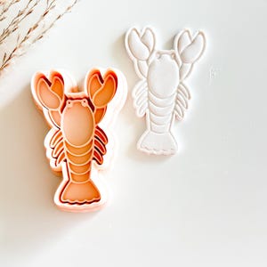 May include: Two lobster-shaped cookie cutters. One is a layered peach-colored cutter, and the other is a white cutter. The cutters are designed with detailed claws, body segments, and tail fins. Ideal for creating themed cookies or fondant decorations.