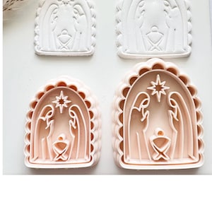 May include: Two light pink plastic cookie cutters and two white embossed clay impressions of a nativity scene. Each piece features an arched frame with a scalloped edge, depicting two figures standing over a central manger with a star above.